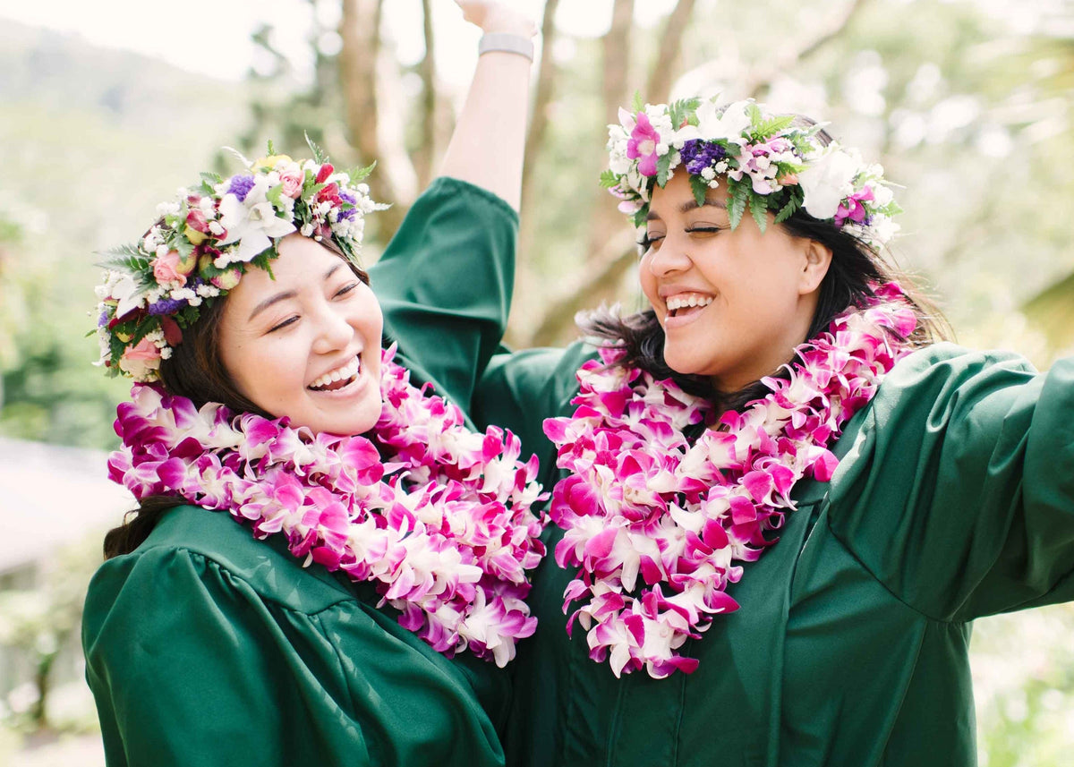 Two people in green graduation gowns with pink leis and flower crowns, standing outdoors with a blurred natural background.